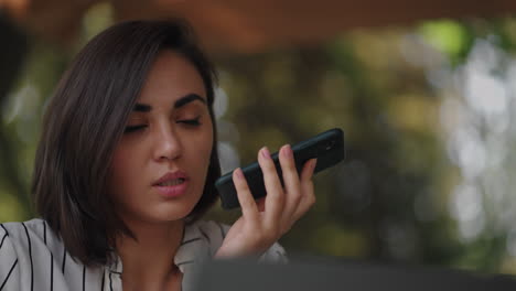 Confident-business-woman-recording-audio-message-on-smartphone-at-street.-Woman-sitting-in-a-summer-cafe-uses-smartphone-voice-recognition-send-voice-message.-speaker-or-leaving-voice-message-concept